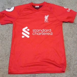 Röd fotbollströja från Liverpool FC - Säljer en röd fotbollströja från Liverpool FC med korta ärmar och klubbens emblem broderat på bröstet. Tröjan har sponsorlogga på framsidan och Premier League-märke på ärmen. Perfekt för alla LFC-fans! YNWA