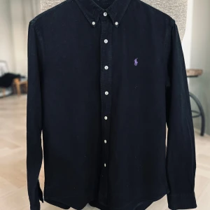 Svart skjorta från Ralph Lauren - Snygg svart skjorta från Ralph Lauren i slim fit. Skjortan har långa ärmar och knappar framtill. Perfekt för en stilren look. Pris kan diskuteras vid snabb affär!