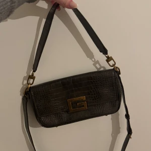 Väska Guess - Snygg brun axelväska från Guess med krokodilpräglat mönster och guldfärgade detaljer. Väskan har en praktisk dragkedja och ett elegant innerfoder med logotyp. 
