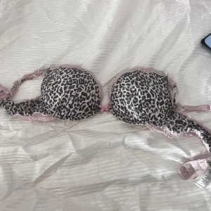 Leopardmönstrad bh med spetsdetaljer - Säljer en snygg leopardmönstrad bh med rosa spetsdetaljer och små rosetter. Bh:n har en bekväm passform och är tillverkad i polyamid och elastan. Perfekt för dig som vill ha något både stilrent och bekvämt.