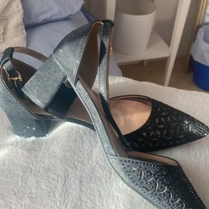 Svarta klackskor från ValenBella - Eleganta svarta klackskor från ValenBella med spetsig tå och glittrig yta. Skorna har en öppen design med remmar och en stabil blockklack. Perfekta för att ge en touch av glamour till din outfit.