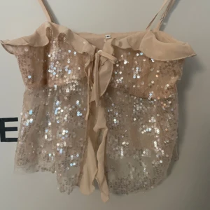 Beige paljettlinne  - Säljer ett glittrande beige linne med paljetter och volangdetaljer. Linnet har smala axelband och en knytning framtill. Perfekt för en festlig kväll!