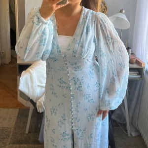 Ljusblå blommig klänning - Charmig ljusblå klänning med blommigt mönster och långa ärmar. Klänningen har en v-ringad design och knappar framtill. Perfekt för vår och sommar.