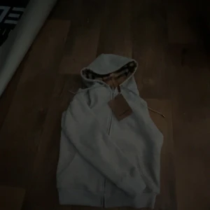 Grå hoodie från Burberry - Säljer en stilren grå hoodie från Burberry med dragkedja och klassiskt rutigt mönster i huvan. Perfekt för en avslappnad och trendig look. Passar bra till både jeans och joggers. Storklek XS och S