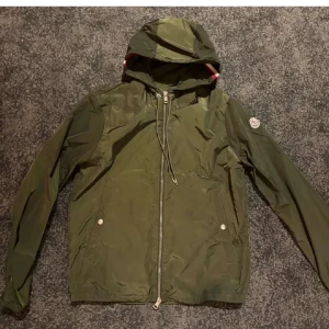 Monclers Olivgröna Windbreaker - Snygg och skön olivgrön windbreaker från Moncler med huva och dragkedja framtill. Jackan har monclers logga på ena armen och en smidig och välfungerande dragkedja. Luvan fungerar toppen då man kan även ändra passform med snörena! Jackan är som perfekt nu till våren, med lite blåst även småkyliga dagar. Några övriga frågor, kontakta mig! 