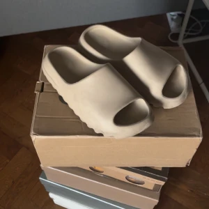 Beige Adidas Yeezy Slide - Säljer ett par beige Adidas Yeezy Slide. Dessa sandaler har en modern och minimalistisk design med öppen tå och en bekväm passform. Perfekta för en avslappnad stil. Fem dotter som 42, men är storlek 44!