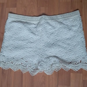 Vita spetsshorts - Säljer en söt vit lågmidjad spetskjol med blommigt mönster. Kjolen har en vacker volangkant och är perfekt för sommarens alla tillfällen. Den är kort och har en elastisk midja för bekväm passform.