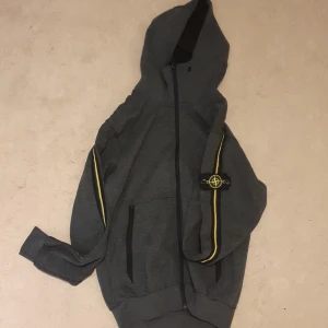 Grå hoodie från Stone Island - Säljer en grå hoodie från Stone Island med dragkedja och huva. Den har en gul och grön rand längs ärmarna och det ikoniska Stone Island-märket på ärmen. Perfekt för en stilren och avslappnad look. Det ni ser på bilden är det ni får😁 ett plagg som är köpt i bulk så jag vet lila lite som ni, men jag har kollat så gott jag kan på alla möjliga sajter och det jag kan kolla stämmer överens med originalet väldigt bra tröja raka o fina sömmar, kvalitets lappar och tyg