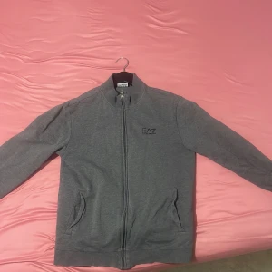 Grå jacka från EA7 Emporio Armani - Snygg grå jacka från EA7 Emporio Armani med dragkedja framtill och två praktiska fickor. Perfekt för en stilren look. Jackan har långa ärmar och en enkel design som passar till mycket.