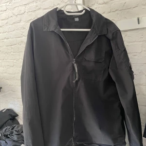 Svart jacka med dragkedja - Säljer en stilren svart jacka från C.P. Company. Jackan har en dragkedja framtill och en ficka på ärmen med en rund detalj. Perfekt för en casual look. Skick 9/10. Nypris 3399. 