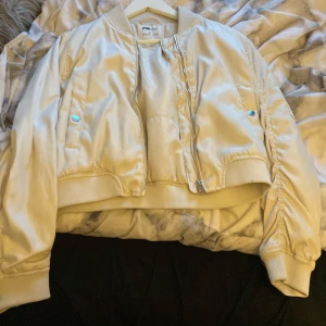 Beige bomberjacka från FS Sister - Snygg beige bomberjacka från FS Sister med dragkedja framtill och ribbade muddar. Jackan har en cool ficka med dragkedja på ärmen och en klassisk bomberstil. Perfekt för en stilren look!