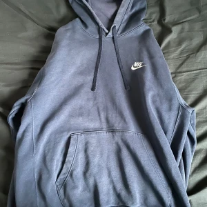 Mörkblå hoodie från Nike - Säljer en snygg mörkblå hoodie från Nike med klassisk logga på bröstet. Tröjan har en stor ficka framtill och justerbar huva med snören. Perfekt för en avslappnad stil.