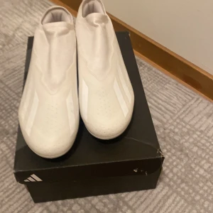 Vita fotbollsskor från Adidas - Snygga vita fotbollsskor från Adidas med en stilren design. Skorna har en strömlinjeformad form utan snörning för en smidig passform. Perfekta för fotbollsplanen med sin moderna look och funktionalitet. Storlek 44 1/2 och oandvärbara.