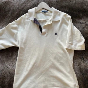 Burberry pike tröja - Säljer en stilren vit pikétröja från Burberry med  krage och korta ärmar. Tröjan har en diskret logga på bröstet och en snygg detalj med rutigt mönster på insidan av kragen. Dm vid intresse 😁