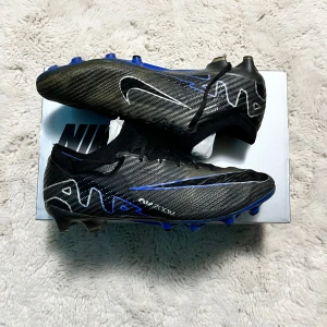 Nike fotbollsskor med blå detaljer - Snygga fotbollsskor från Nike i svart med blå och vita detaljer. Skorna har en modern design med snörning och en rund tå. Perfekta för fotbollsplanen med sin slitstarka sula och stilrena look. Skick: 8/10 (priset kan diskuteras?)