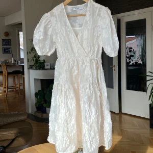 Vit blommig klänning med knytband - Charmig vit klänning med blommigt mönster och knytband i midjan. Klänningen har korta puffärmar och en omlott-design som ger en romantisk känsla. Perfekt för sommarens festligheter.