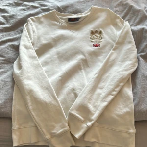 Morris Vit sweatshirt med broderad logga - Säljer en stilren vit sweatshirt med en broderad logga och brittisk flagga på bröstet. Tröjan har långa ärmar och ribbade muddar vid ärmslut och nederkant. Perfekt för en avslappnad stil.