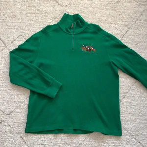 Grön tröja med dragkedja och broderi - Snygg grön tröja med halv dragkedja och ett broderat motiv av polospelare på bröstet. Perfekt för en sportig look med stil. Tröjan har långa ärmar och en hög krage.