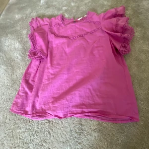 Rosa blus med volangdetaljer - Charmig rosa blus med volangdetaljer och korta ärmar. Blusen har dekorativa spetsdetaljer vid halsen och ärmarna, vilket ger en romantisk touch. Perfekt för en somrig look. skriv för flera bilder🥰