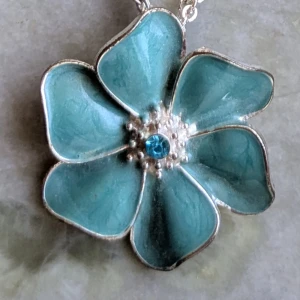 Blått blomformat halsband med kedja - Charmigt halsband med en stor blomma. Blomman har en glittrande blå sten i mitten och hänger på en elegant kedja. Perfekt för att ge en färgklick till din outfit.