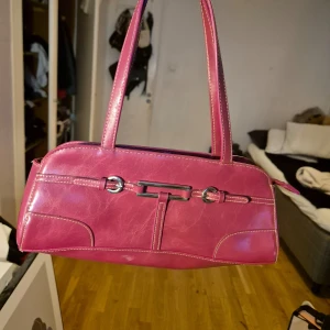 Rosa handväska med silverdetaljer - Snygg rosa handväska i syntetmaterial med silverdetaljer på framsidan. Väskan har två långa handtag och en stilren design. Perfekt för att ge en färgklick till din outfit.