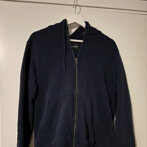 Mörkblå hoodie från Jack & Jones - Säljer en stilren mörkblå hoodie från Jack & Jones. Den har en dragkedja framtill och en praktisk huva med snören. Perfekt för en avslappnad look.