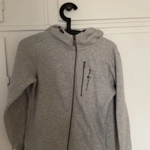 Grå hoodie med dragkedja - Säljer en grå hoodie från Peak Performance med dragkedja och huva. Perfekt för en avslappnad stil. Den har en diskret logga på bröstet och är tillverkad i ett mjukt material.