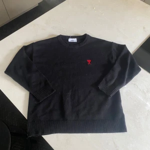 Svart tröja från Ami Paris - Säljer en stilren svart tröja från Ami Paris med ett rött broderat hjärta och bokstaven A på bröstet. Tröjan har långa ärmar och ribbade muddar vid ärmslut och nederkant. Perfekt för en minimalistisk look.