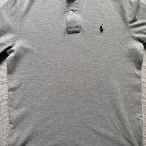 Grå pikétröja från Ralph Lauren - Säljer en stilren grå pikétröja från Ralph Lauren med klassisk krage och knappar framtill. Tröjan har en broderad logotyp på bröstet. Perfekt för en avslappnad men ändå snygg look.