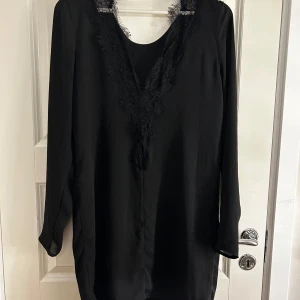 Svart klänning med spetsdetaljer - Elegant svart klänning med långa ärmar och v-ringad spetsdetalj framtill. Perfekt för en stilren look.