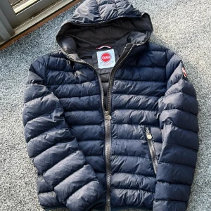 Mörkblå pufferjacka från Colmar - Säljer en snygg mörkblå pufferjacka från Colmar med huva och dragkedja. Jackan har långa ärmar och praktiska fickor med dragkedjor. Perfekt för kyliga dagar!