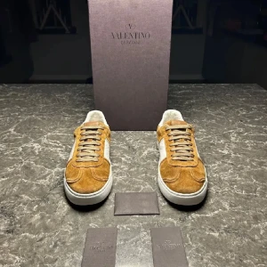 Bruna mocka sneakers från Valentino - Snygga bruna sneakers i mocka från Valentino med vita detaljer och snörning. Skorna har en unik sula med ett subtilt mönster och en logotypdetalj. Perfekta för en stilren look. Storlek 41 men passar mig som brukar ha 42/43. Innersulan är 27cm. OBS EN NIT SAKNAS‼️