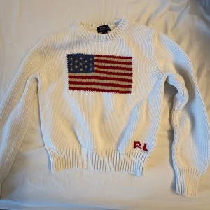 Vit stickad tröja från Ralph Lauren - Säljer en vit stickad tröja från Ralph Lauren med en stor amerikansk flagga på framsidan. Tröjan har långa ärmar och en rund halsringning. Perfekt för en stilren och klassisk look.