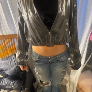 Silverfärgad glittrig jacka - Snygg silverfärgad glittrig jacka med dragkedja framtill. Jackan har en croppad passform och långa ärmar, perfekt för en kväll ute. Den glänsande ytan ger en festlig touch.