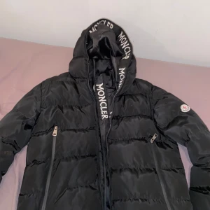 Svart pufferjacka från Moncler - Säljer en snygg svart pufferjacka från Moncler. Jackan har en stilren design med dragkedja och Moncler-logga på ärmen. Perfekt för kyliga dagar med sin vadderade och quiltade stil. Huvan är lite skadad men det syns inte och man kan sy den enkelt