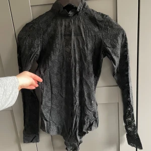 Svart spetsbody - Svart spetsbody med långa ärmar och hög hals. Bodyn har knäppning i grenen och är lite öppen i ryggen. 