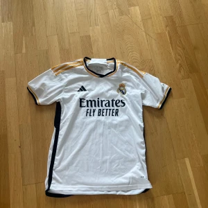 Real Madrid tröja - Säljer en vit fotbollströja från Adidas med korta ärmar och svarta detaljer. Tröjan har guldfärgade ränder på axlarna och klubbmärke på bröstet. Perfekt för fotbollsfans!