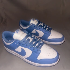 Nike Dunk i blått och vitt - Snygga Nike Dunk sneakers i en fräsch kombination av blått och vitt. Skorna har en klassisk design med snörning och perforerade detaljer på tån för extra ventilation. Perfekta för en stilren och sportig look.