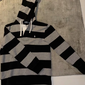 Randig hoodie från Polo Ralph Lauren - Säljer en snygg randig hoodie från Polo Ralph Lauren i svart och grått. Tröjan har en klassisk design med huva och dragsko samt långa ärmar. Perfekt för en avslappnad stil. Priset är diskuterbart!