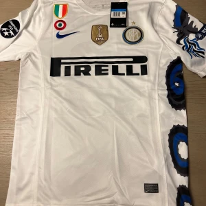 Vit Inter Milan fotbollströja - Säljer en vit Inter Milan fotbollströja från Nike med Pirelli-logga på framsidan. Tröjan har korta ärmar och ett unikt mönster i blått och svart längs sidorna. Den har även märken för FIFA och Champions League. Namnet 'Sneijder' och nummer 10 pryder baksidan.