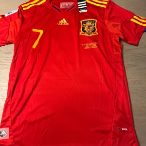 Röd fotbollströja från Adidas - Säljer en röd fotbollströja från Adidas med gula detaljer och nummer 7 på framsidan. Tröjan har korta ärmar och ett emblem på bröstet. Perfekt för fotbollsfans!