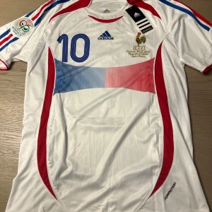 Vit fotbollströja med nummer 10 - Säljer en vit fotbollströja från Adidas med nummer 10 och namn på ryggen. Tröjan har korta ärmar och detaljer i blått och rött. Perfekt för fotbollsfans som vill ha en klassisk look.