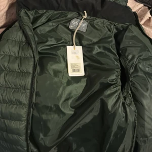 Grön quiltad väst från Lyle & Scott - Säljer en snygg grön quiltad väst från Lyle & Scott. Jackan har en lätt och bekväm design med dragkedja och en liten logotyp på bröstet. Perfekt för kyligare dagar.