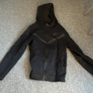Nike tech 137-146 - Säljer en svart Nike tech  med dragkedja och huva. Tröjan har långa ärmar och ett diskret Nike-logotyp på bröstet. Perfekt för en sportig look eller avslappnade dagar. Priset är inte spikat i sten. ny pris=1100 
