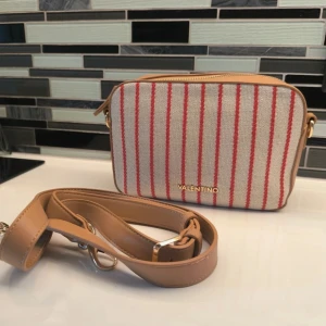 Randig axelväska från Valentino - Snygg axelväska från Valentino med röd- och vitrandigt mönster. Väskan har en justerbar axelrem i beige och en praktisk dragkedja upptill. Perfekt för att ge din outfit en stilren touch.