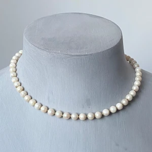 Pärlhalsband med dekorativt lås - Elegant pärlhalsband med vita pärlor och ett dekorativt lås i silverfärg. Perfekt för att ge en klassisk touch till din outfit. Stämpel silver  42 cm långt med knut mellan varje pärla  Skicket är okej, tecken på halsbandets ålder finns på några av pärlorna