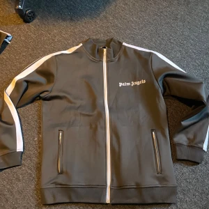 Svart träningsjacka från Palm Angels och byxor  - Snygg svart träningsjacka från Palm Angels med vita ränder längs ärmarna och dragkedja framtill. Jackan har två fickor med dragkedja och en stilren logga på bröstet. Perfekt för en sportig look.