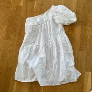 Vit klänning, one shoulder - Säljer en söt vit klänning från Boohoo med en offshoulder-design och kort ärm. Klänningen har fina detaljer och en luftig passform, perfekt för varma dagar. Den är gjord i ett lätt och bekvämt material. Aldrig använd!