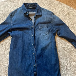 Blå jeansskjorta från Replay - Snygg blå jeansskjorta från Replay med en klassisk krage och knappar framtill. Skjortan har en bröstficka och långa ärmar, perfekt för en avslappnad stil. Märket Replay är broderat på fickan. Passar perfekt nu till sommaren och går att styla med mycket, även en skön passform i storlek S