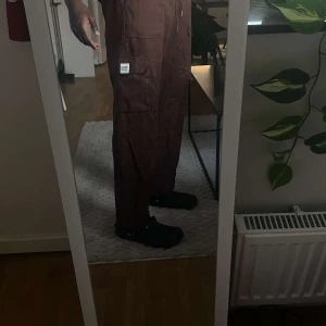 Bruna cargopants med fickor - Säljer ett par bruna cargopants med flera stora fickor på sidorna och snörning i midjan. Byxorna har en loose passform och raka ben, perfekta för en chill och avslappnad stil. Materialet är bomull och de har en patchdetalj på ena fickan.